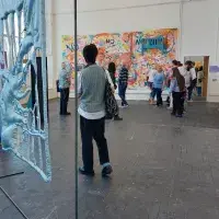 Exhibition shot showing blue fabric handing over a metal frame in the foreground to the right. A male presenting person walks towards a group of people in the background.