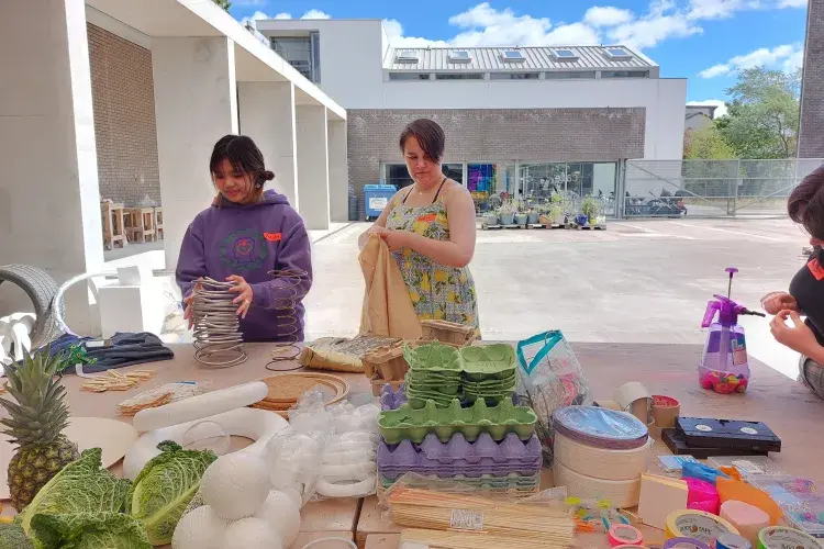 A colourful collection of objects are gathered on a table and a couple of people are picking up objects to play with them.
